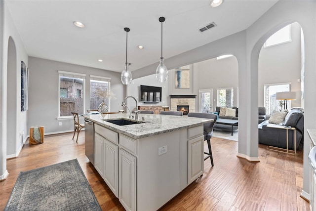 kitchen with sink, hardwood / wood-style flooring, dishwasher, light stone counters, and an island with sink