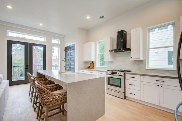 kitchen with wall chimney range hood, sink, white cabinets, a center island with sink, and gas range