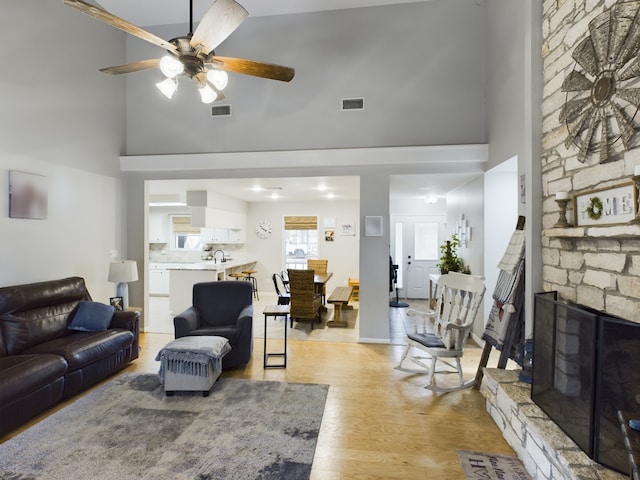 living room featuring a towering ceiling, light hardwood / wood-style flooring, a stone fireplace, and ceiling fan