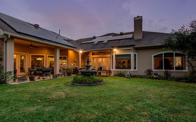 back house at dusk with ceiling fan, a lawn, and solar panels