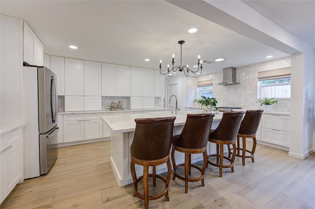 kitchen featuring wall chimney exhaust hood, decorative light fixtures, stainless steel refrigerator, a kitchen island with sink, and white cabinets