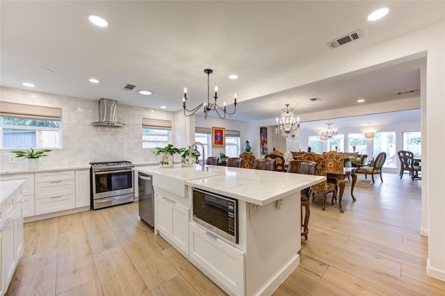 kitchen featuring wall chimney exhaust hood, appliances with stainless steel finishes, a kitchen island with sink, and white cabinets