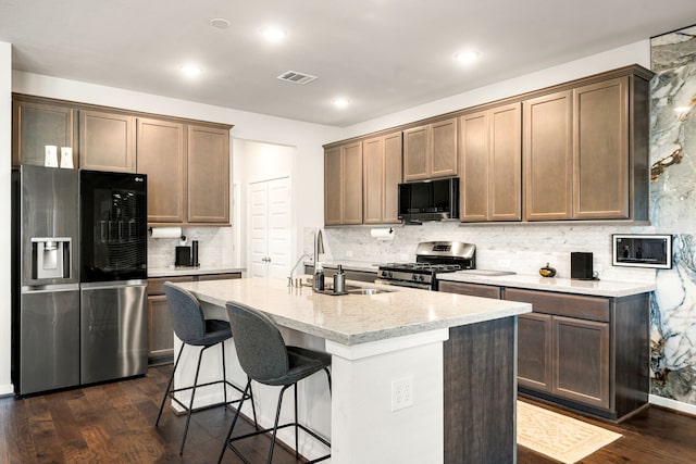 kitchen with dark wood-type flooring, sink, light stone counters, a center island with sink, and stainless steel appliances