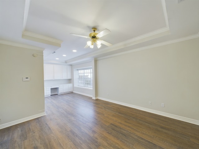 unfurnished living room with ceiling fan, a tray ceiling, built in desk, ornamental molding, and dark hardwood / wood-style flooring