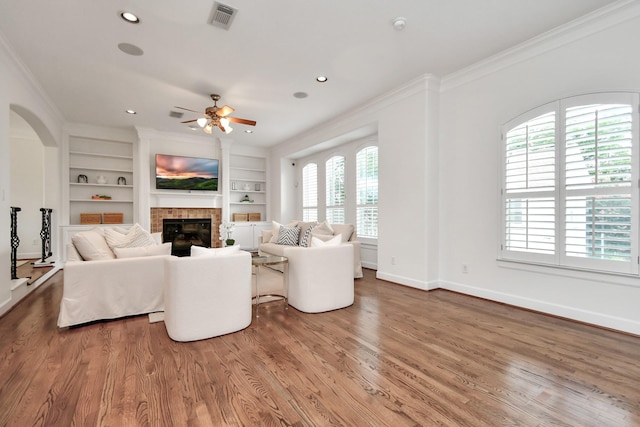 living room with crown molding, wood-type flooring, a brick fireplace, built in features, and ceiling fan