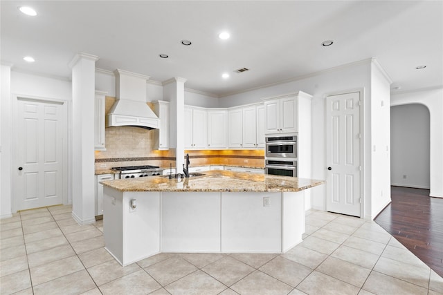 kitchen featuring white cabinetry, light stone counters, a center island with sink, and premium range hood