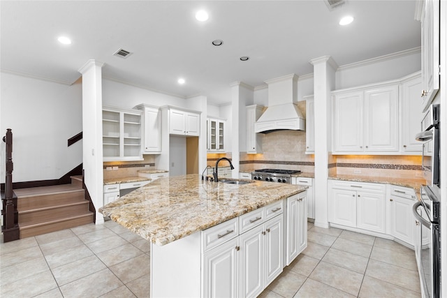 kitchen featuring premium range hood, white cabinetry, sink, light stone counters, and a center island with sink