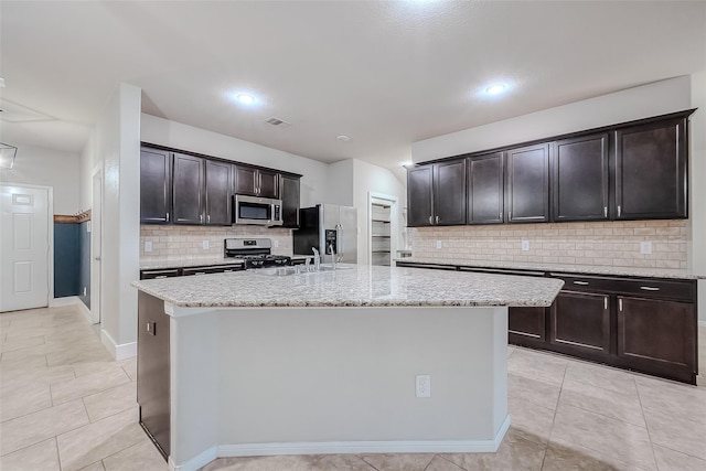 kitchen featuring appliances with stainless steel finishes, an island with sink, light tile patterned floors, light stone counters, and dark brown cabinetry