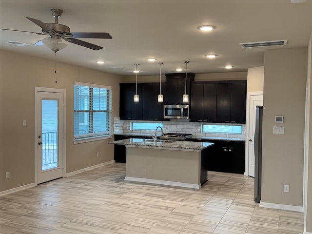 kitchen featuring light stone countertops, a kitchen island with sink, decorative backsplash, and decorative light fixtures