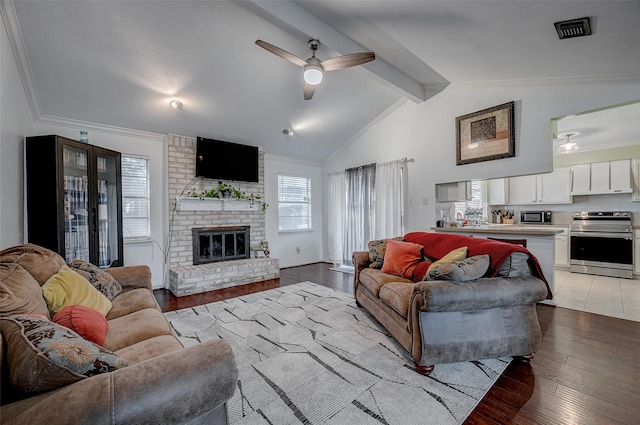 living room featuring ceiling fan, ornamental molding, lofted ceiling with beams, a brick fireplace, and light wood-type flooring