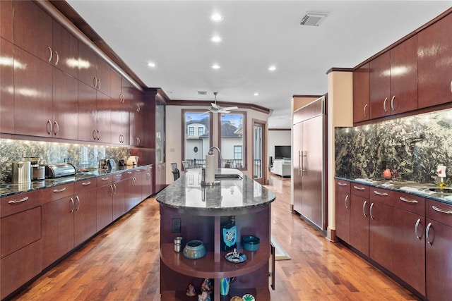 kitchen with sink, crown molding, dark stone countertops, paneled fridge, and a center island with sink