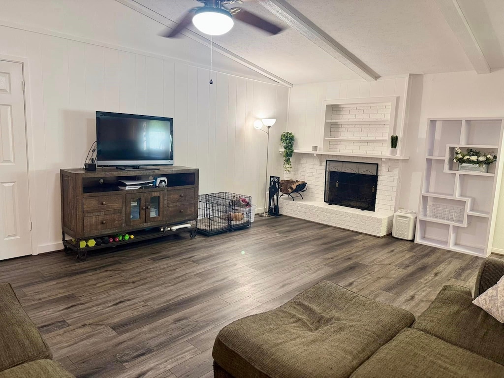 living room with beamed ceiling, a brick fireplace, wood finished floors, and a ceiling fan