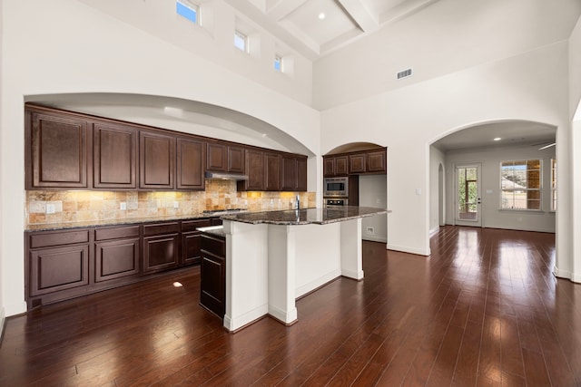 kitchen with appliances with stainless steel finishes, dark hardwood / wood-style flooring, a kitchen island with sink, and dark stone countertops