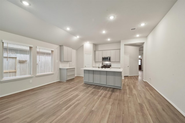 kitchen with lofted ceiling, gray cabinets, appliances with stainless steel finishes, light hardwood / wood-style floors, and a center island with sink