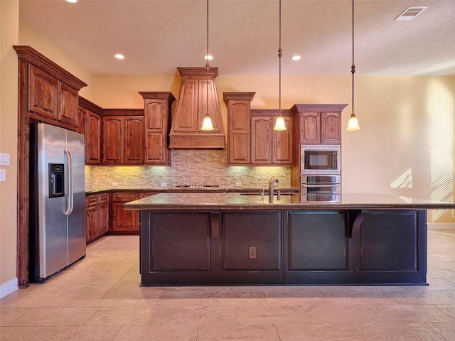 kitchen featuring premium range hood, hanging light fixtures, appliances with stainless steel finishes, an island with sink, and light stone countertops