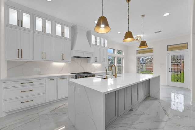 kitchen with sink, custom exhaust hood, white cabinetry, hanging light fixtures, and an island with sink