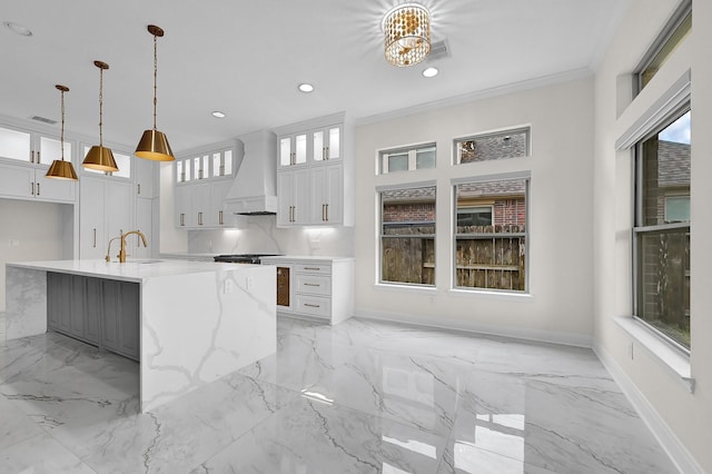 kitchen with premium range hood, white cabinetry, light stone counters, hanging light fixtures, and a kitchen island with sink