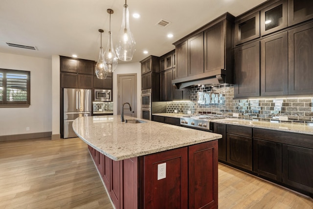 kitchen with premium range hood, decorative light fixtures, a kitchen island with sink, stainless steel appliances, and light stone countertops