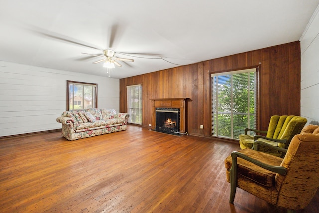 living room with wooden walls, a warm lit fireplace, wood finished floors, and ceiling fan