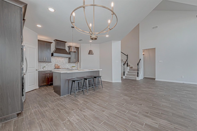 kitchen featuring premium range hood, a kitchen island with sink, hanging light fixtures, a notable chandelier, and decorative backsplash