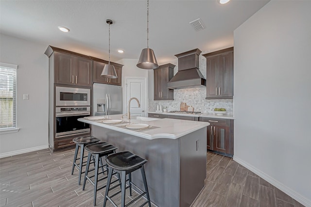 kitchen with pendant lighting, a kitchen island with sink, backsplash, stainless steel appliances, and custom range hood