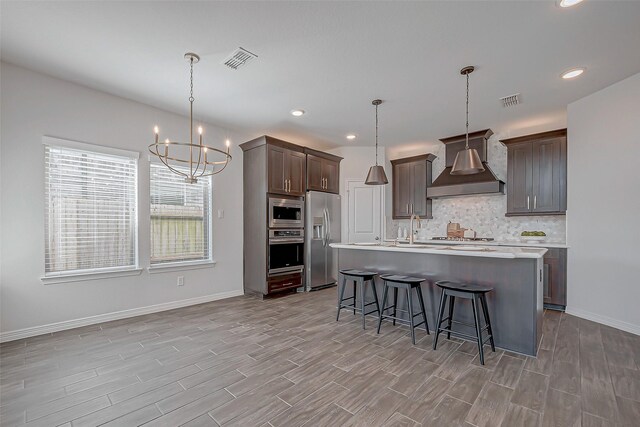 kitchen featuring hanging light fixtures, a center island with sink, wall chimney range hood, and appliances with stainless steel finishes