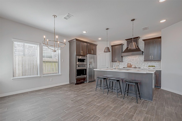 kitchen featuring a center island with sink, appliances with stainless steel finishes, wall chimney exhaust hood, and decorative light fixtures