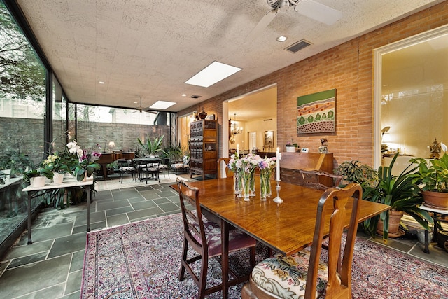 dining area with ceiling fan with notable chandelier, a skylight, and brick wall