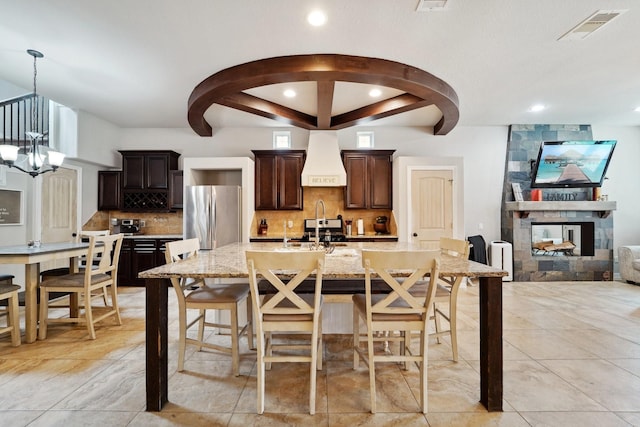 kitchen featuring premium range hood, pendant lighting, an island with sink, stainless steel fridge, and a kitchen bar