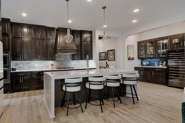 kitchen featuring dark brown cabinetry, custom exhaust hood, hanging light fixtures, an island with sink, and stainless steel gas stovetop