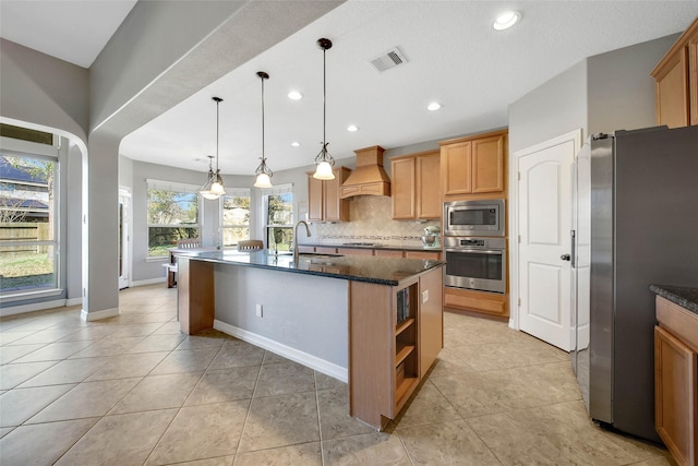 kitchen featuring sink, appliances with stainless steel finishes, a kitchen island with sink, custom range hood, and decorative light fixtures