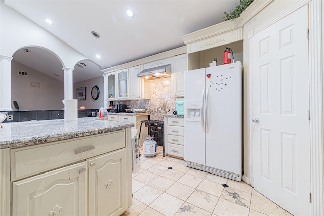 kitchen featuring vaulted ceiling, decorative columns, white refrigerator with ice dispenser, light stone countertops, and wall chimney exhaust hood