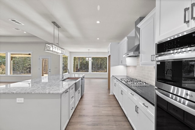 kitchen featuring appliances with stainless steel finishes, white cabinetry, hanging light fixtures, a large island with sink, and wall chimney exhaust hood