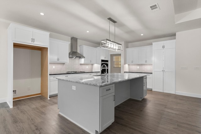 kitchen featuring light stone counters, wall chimney range hood, an island with sink, stainless steel appliances, and white cabinets