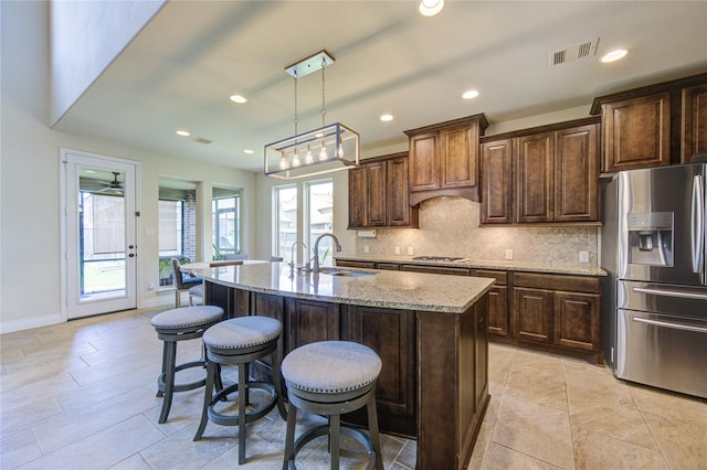 kitchen with sink, decorative backsplash, light stone counters, stainless steel appliances, and a center island with sink