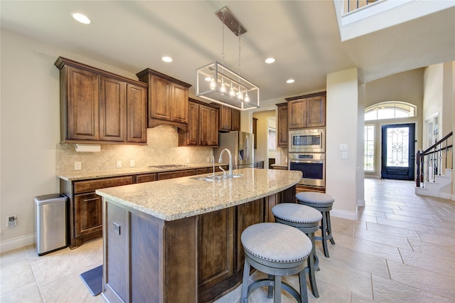 kitchen featuring appliances with stainless steel finishes, a kitchen island with sink, hanging light fixtures, light stone counters, and decorative backsplash