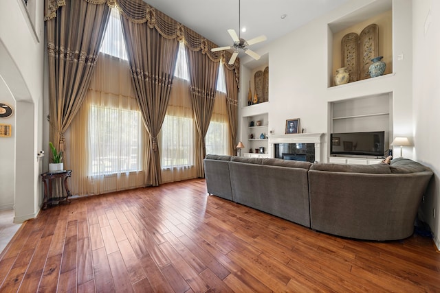 unfurnished living room with built in shelves, wood-type flooring, ceiling fan, and a high ceiling