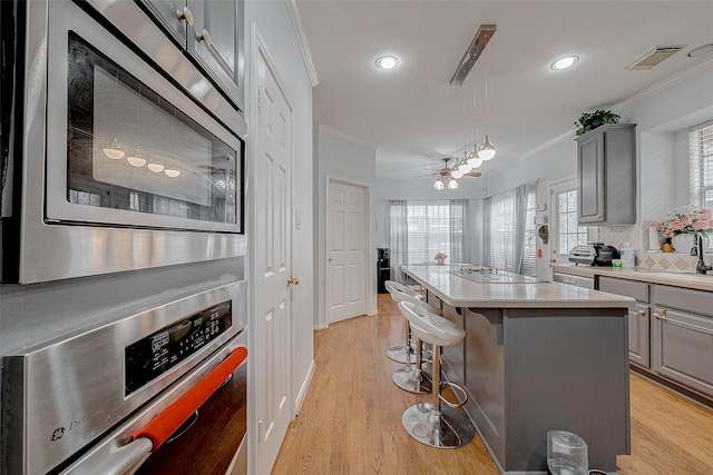 kitchen with gray cabinetry, ornamental molding, a kitchen island with sink, stainless steel appliances, and light hardwood / wood-style flooring