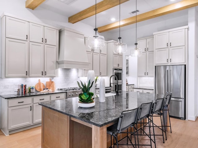 kitchen with hanging light fixtures, stainless steel appliances, a center island with sink, custom exhaust hood, and dark stone counters