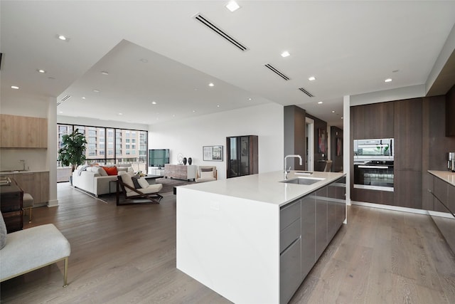 kitchen with a kitchen island with sink, sink, stainless steel double oven, and light wood-type flooring