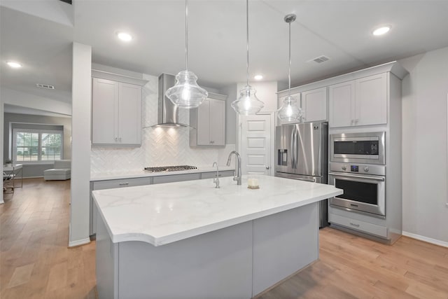 kitchen featuring an island with sink, appliances with stainless steel finishes, wall chimney range hood, and decorative light fixtures