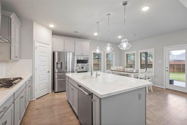 kitchen featuring appliances with stainless steel finishes, decorative backsplash, hanging light fixtures, light stone countertops, and a center island with sink