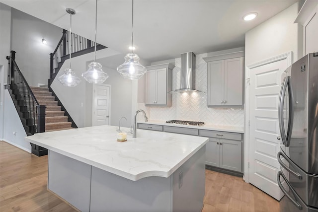 kitchen with gray cabinets, hanging light fixtures, a kitchen island with sink, stainless steel appliances, and wall chimney exhaust hood