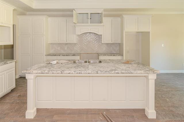 kitchen featuring a kitchen island with sink, backsplash, white cabinetry, light stone counters, and ornamental molding