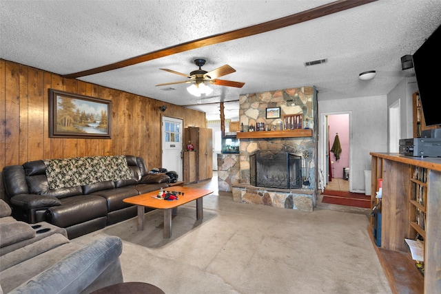 living room with ceiling fan, beam ceiling, a textured ceiling, a stone fireplace, and wood walls