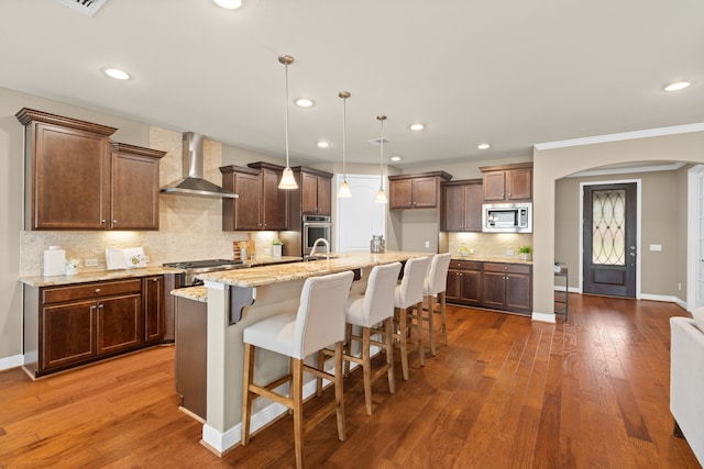 kitchen with stainless steel microwave, wall chimney range hood, recessed lighting, a center island with sink, and arched walkways