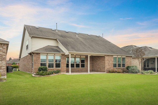 back of house at dusk featuring central AC, a yard, roof with shingles, brick siding, and a patio area