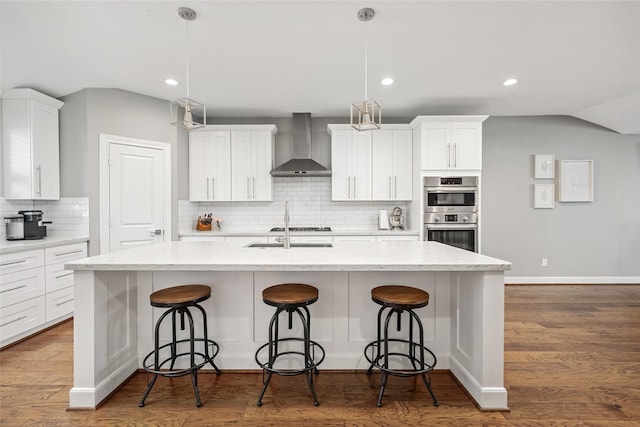 kitchen with pendant lighting, wall chimney range hood, an island with sink, and appliances with stainless steel finishes