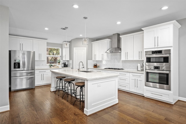 kitchen with stainless steel appliances, a sink, white cabinets, wall chimney exhaust hood, and a center island with sink