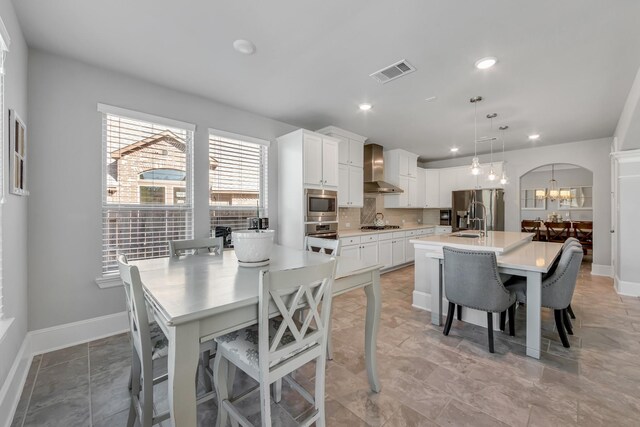 kitchen featuring a center island with sink, hanging light fixtures, wall chimney range hood, stainless steel appliances, and white cabinets